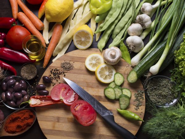 Photo from directly above of a kitchen knife and sliced vegetables on a cutting board and variation of vegetables and spices around.