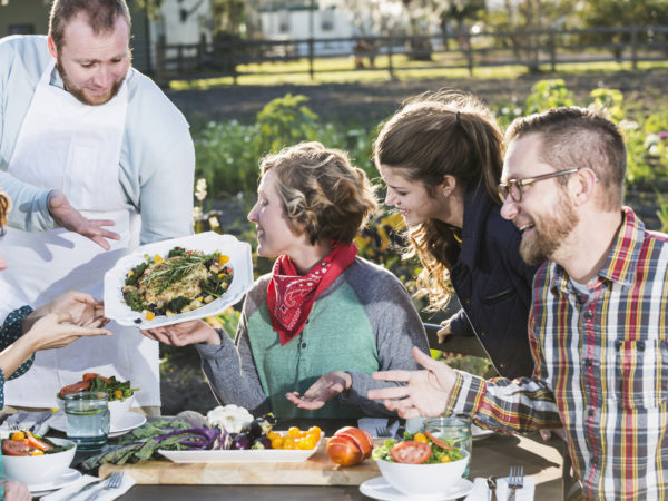 A group of people sitting outdoors at a dining table, being served locally grown food. The chef or waiter is standing, presenting a dish of beautifully prepared vegetables. This image represents the farm-to- table-or-farm to fork movement.