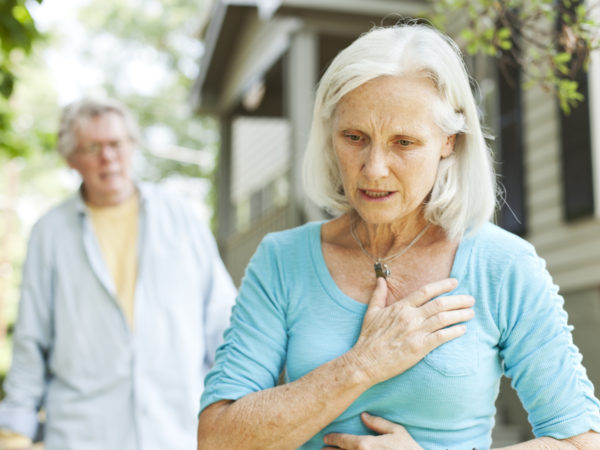 A senior woman experiencing chest pain. A senior man is in the background.