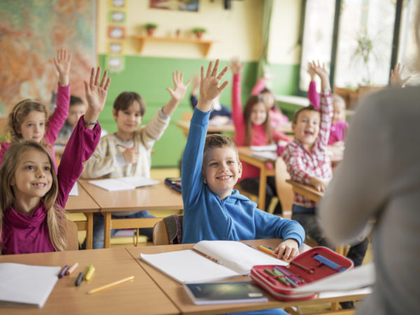 Large group of school children raising their hands ready to answer the question.