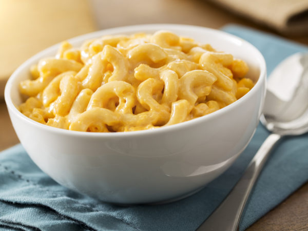 A white bowl of the best baked homemade macaroni and cheese on a table with a blue napkin. The recipe is made with elbow pasta and cheddar cheese. The shot is vertical and has selective focus. Macaroni and cheese is a American comfort food or soul food.