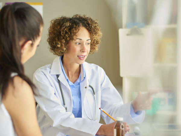 A female doctor sits at her desk and explains diagnosis to a female patient . She is wearing a white coat and stethoscope and is referring to a computer screen whilst making notes.