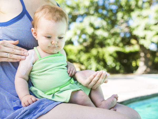 A close up of a mom and her smiling baby girl sit next to a pool, the mom applying sunblock to her daughters skin to protect from the suns harmful U.V. rays. The child smiles, patiently letting her put on the lotion. Horizontal image with copy space.