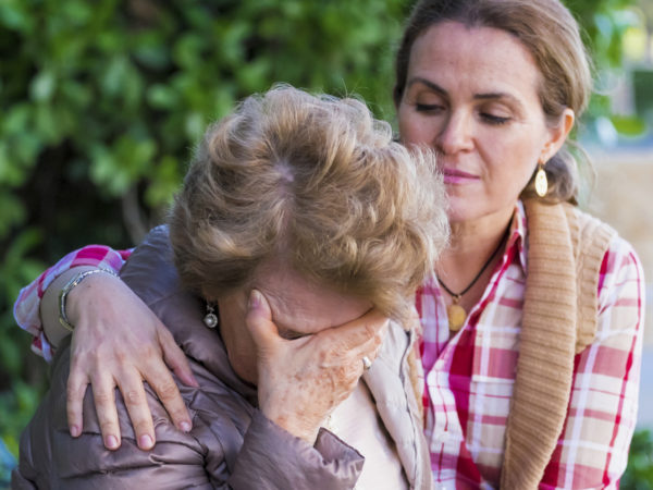Senior woman with her hands on her head looking down sitting in a park bench with her daughter consoling her
