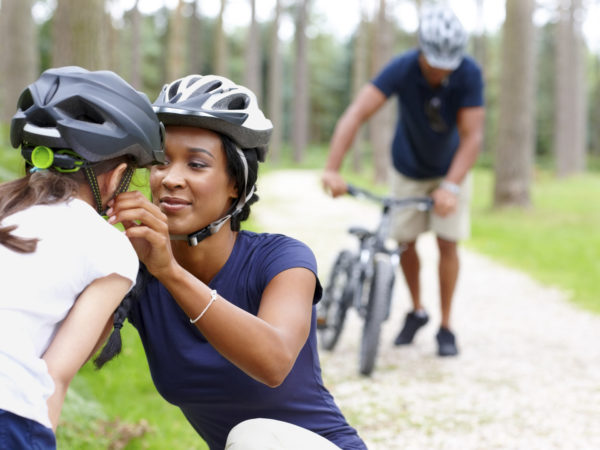 Caring young mother helping daughter put on helmet with father in background - Outdoors in country side