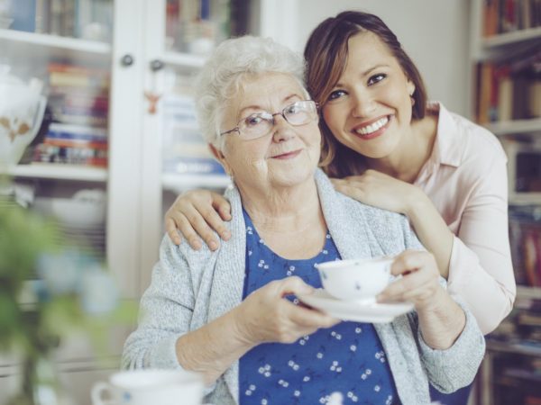 Social worker is visiting a senior woman in her own apartment. They are celebrating the elderly lady's birthday. The kind nurse is hugging the senior woman over her shoulder.