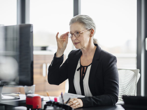 Portrait of a senior businesswoman sitting on her desk in a modern office and. She's working on a computer and wearing glasses and has silver hair.