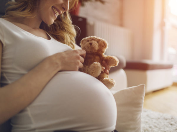 Pregnant Woman Holding Teddy Bear and smiling, sitting on the floor in her living room. Wearing white T-shirt . Woman has brown curly hair and beautiful smile. Sunlight in background.