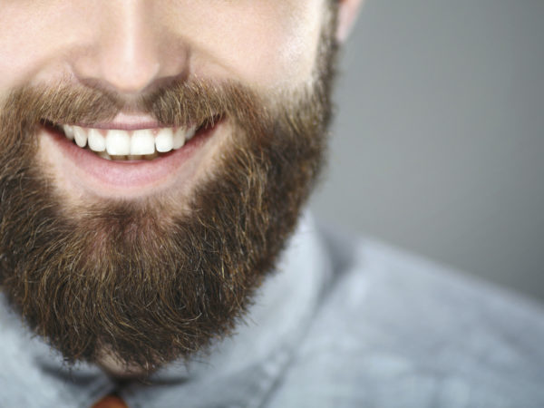 Closeup portrait of smiling unrecognizable bearde man. He's facing the camera and smiling with perfect set of teeth. Wearing light blue smart casual shirt. Shot oer gray background.
